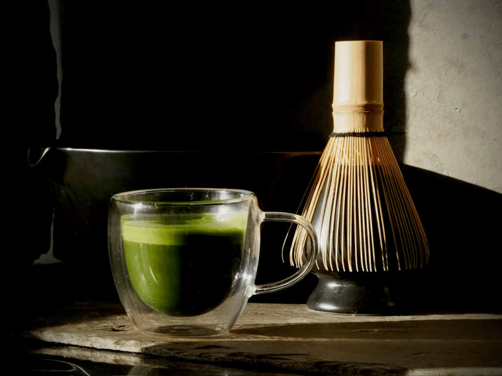 The image depicts a matcha whisk and a teacup containing matcha. At right, a traditional bamboo "chasen" matcha whisk with...